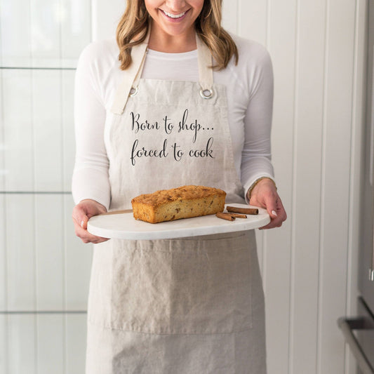 a woman holding a plate with a loaf of bread on it wearing an apron that says born to shop...forced to cook