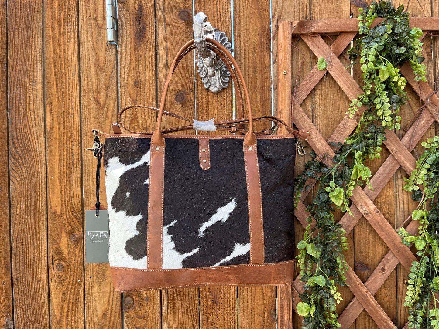 a black and white cow print purse with brown straps hanging on a wooden wall